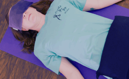 Woman reclined in savasana, the final resting pose after a yoga class. She has a purple eye pillow on her eyes, a purple sandbag on her hips, and is reclined on a purple yoga mat.